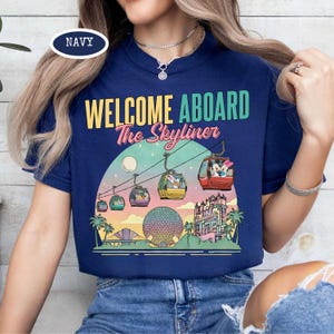 May include: Navy blue t-shirt with the text "WELCOME ABOARD The Skyliner" above a graphic of a gondola ride with cartoon characters. The design includes a colorful sunset and various landmarks.
