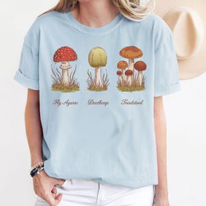 May include: Light blue t-shirt with a graphic of three different types of mushrooms. The mushrooms are labeled "Fly Agaric", "Deathcap", and "Toadstool".