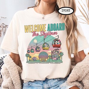 May include: Ivory-colored t-shirt with a colorful graphic featuring the text "WELCOME ABOARD The Skyliner." The design includes gondolas, a moon, palm trees, and buildings, creating a travel theme.