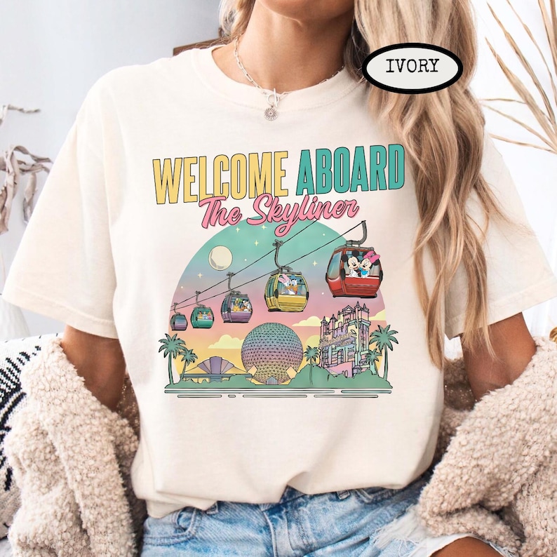 May include: Ivory-colored t-shirt with a colorful graphic featuring the text "WELCOME ABOARD The Skyliner." The design includes gondolas, a moon, palm trees, and buildings, creating a travel theme.