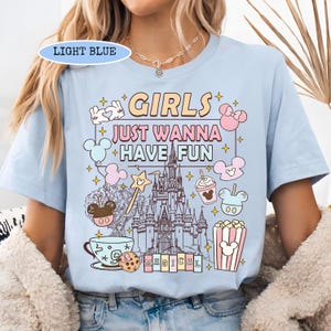 May include: Light blue t-shirt with the text "GIRLS JUST WANNA HAVE FUN" and a castle graphic. The design includes cartoon images of Mickey Mouse, popcorn, and a coffee cup. The shirt is a light blue color.