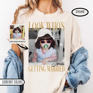 May include: An ivory short-sleeve t-shirt featuring a custom photo print of a child wearing a white bucket hat, red sunglasses, and a pacifier. The text reads LOOK WHO'S GETTING MARRIED and includes the name Sophia in a script font.