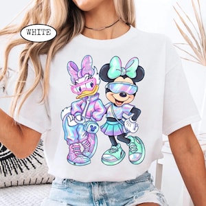 May include: White t-shirt featuring a colorful graphic of Daisy Duck and Minnie Mouse wearing sunglasses and stylish outfits. The word "WHITE" is printed on the upper left of the shirt. The design has a pastel, iridescent color scheme.