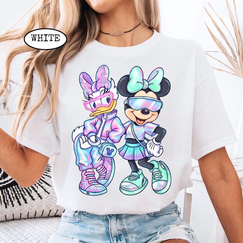 May include: White t-shirt featuring a colorful graphic of Daisy Duck and Minnie Mouse wearing sunglasses and stylish outfits. The word "WHITE" is printed on the upper left of the shirt. The design has a pastel, iridescent color scheme.