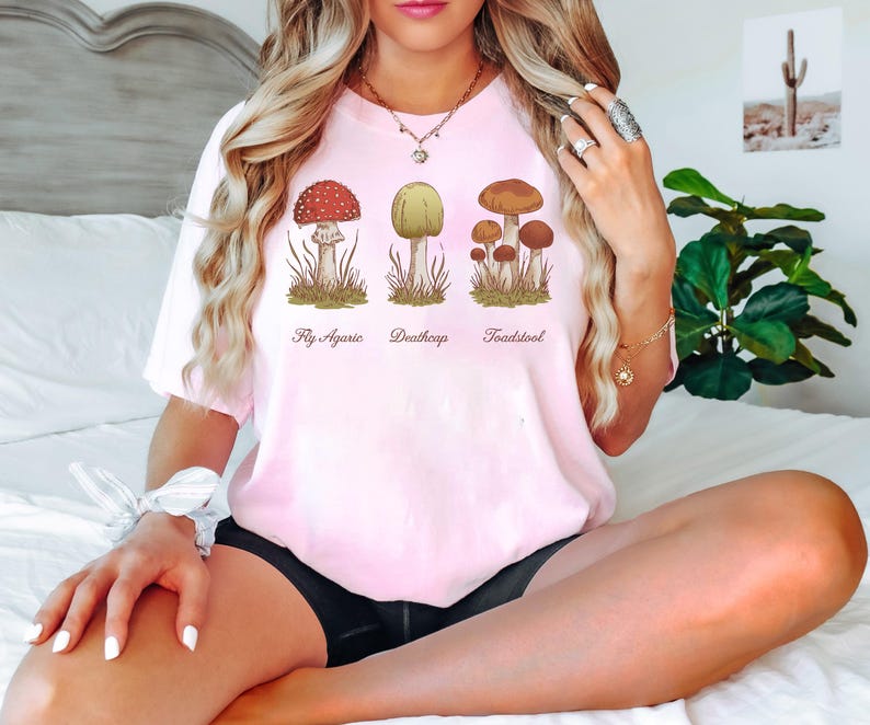 May include: Pink t-shirt with a graphic of three different types of mushrooms. The mushrooms are labeled "Fly Agaric", "Deathcap", and "Toadstool".