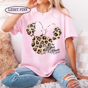 May include: Pale pink t-shirt with a Minnie Mouse design. The design features a leopard print bow and ears, with the text "Minnie Mouse xoxo" in black script. The shirt is short-sleeved and made of a soft material.