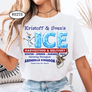 Op de afbeelding: Wit T-shirt met de blauwe tekst "Kristoff & Sven's ICE", met Olaf- en Sven-graphics. Het shirt bevat ook tekst over het oogsten en bezorgen van ijs in het hele koninkrijk Arendelle.