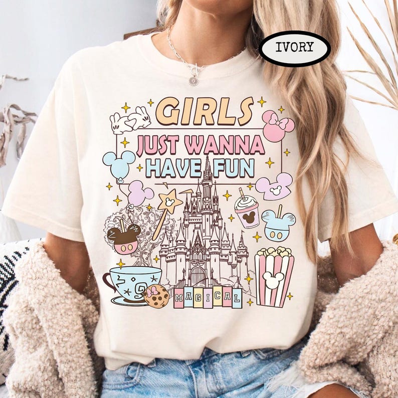 May include: Ivory-colored t-shirt with the text "GIRLS JUST WANNA HAVE FUN" in pastel colors, along with cartoon illustrations of a castle, Mickey Mouse, popcorn, and other magical elements.