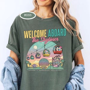May include: Moss green t-shirt with the text "WELCOME ABOARD The Skyliner" in yellow, teal, and pink. The graphic features a sunset scene with gondolas, a sphere, and a castle. The shirt is a comfortable, casual style.