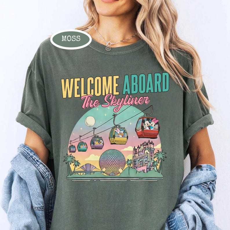 May include: Moss green t-shirt with the text "WELCOME ABOARD The Skyliner" in yellow, teal, and pink. The graphic features a sunset scene with gondolas, a sphere, and a castle. The shirt is a comfortable, casual style.