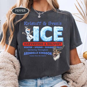 Op de afbeelding: Donkergrijs T-shirt met de blauwe tekst "Kristoff & Sven's ICE", met cartoonafbeeldingen van Olaf en Sven. Het shirt bevat ook de tekst "Harvesting & Delivery" en "Arendelle Kingdom".