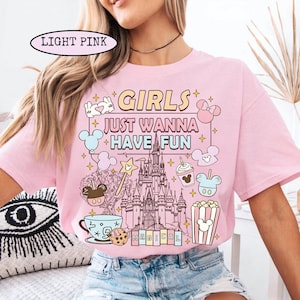 May include: Light pink t-shirt with the text "GIRLS JUST WANNA HAVE FUN" and a graphic of a castle, Mickey Mouse, and other Disney-themed elements. The shirt has short sleeves and a crew neck.