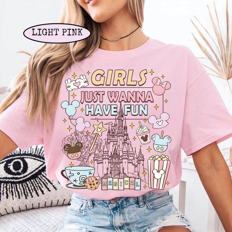 May include: Light pink t-shirt with the text "GIRLS JUST WANNA HAVE FUN" and a graphic of a castle, Mickey Mouse, and other Disney-themed elements. The shirt has short sleeves and a crew neck.