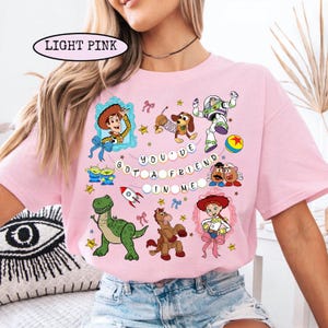 May include: Light pink t-shirt featuring cartoon characters from a popular animated movie. The shirt has the text "YOU'VE GOT A FRIEND IN ME" and images of Woody, Buzz Lightyear, and other characters. The shirt is a crew neck style.