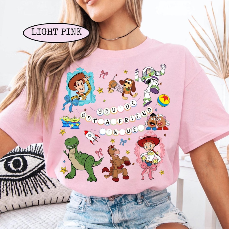 May include: Light pink t-shirt featuring cartoon characters from a popular animated movie. The shirt has the text "YOU'VE GOT A FRIEND IN ME" and images of Woody, Buzz Lightyear, and other characters. The shirt is a crew neck style.