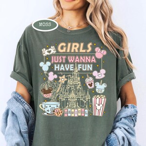 May include: Moss green t-shirt with the text "GIRLS JUST WANNA HAVE FUN" in a colorful, whimsical font. The design includes cartoon illustrations of a castle, Mickey Mouse, popcorn, and other fun elements. The shirt has short sleeves.