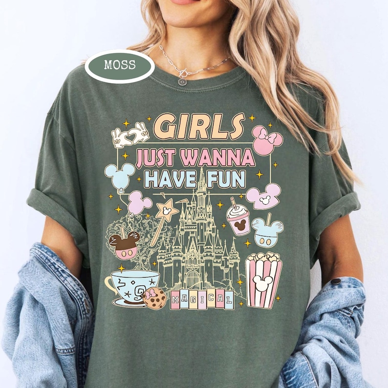 May include: Moss green t-shirt with the text "GIRLS JUST WANNA HAVE FUN" in a colorful, whimsical font. The design includes cartoon illustrations of a castle, Mickey Mouse, popcorn, and other fun elements. The shirt has short sleeves.