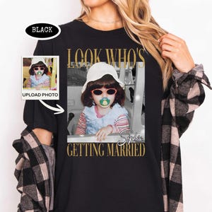 May include: A black t-shirt featuring a graphic print of a child wearing a white bucket hat, red-rimmed sunglasses, and a pacifier. The text reads LOOK WHO'S GETTING MARRIED with the name Sophia. A small inset box shows the original photo upload.