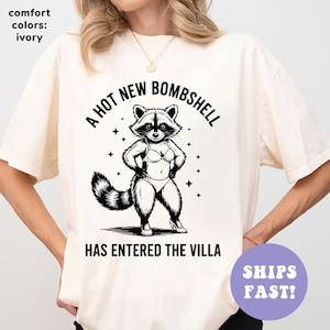 May include: Ivory-colored t-shirt with a black graphic of a raccoon in a bikini, with the text "A HOT NEW BOMBSHELL HAS ENTERED THE VILLA." The shirt also has the text "SHIPS FAST!"