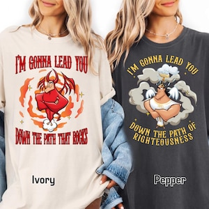 May include: Ivory and pepper-colored t-shirts with graphic designs. The ivory shirt features a red devil illustration with the text "I'M GONNA LEAD YOU DOWN THE PATH THAT ROCKS." The pepper shirt displays an angel with the text "I'M GONNA LEAD YOU DOWN THE PATH OF RIGHTEOUSNESS."