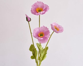 May include: A close-up of artificial pink poppy flowers with yellow centers and green stems and leaves. The flowers are in various stages of bloom, with some fully open and others still in bud form. The background is plain white.