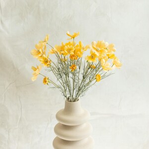 Puede incluir: Un ramo de flores de cosmos amarillas en un jarrón beige con anillos apilados. Las flores tienen pétalos delicados y están dispuestas con tallos verdes. El jarrón está sobre una superficie blanca contra un fondo blanco.