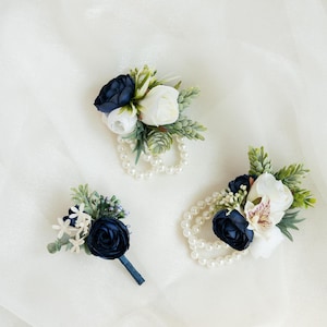 Puede incluir: Tres boutonnières florales azul marino y blancas con detalles de perlas. Las boutonnières están dispuestas sobre un fondo blanco.