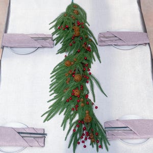 May include: A festive Christmas table setting with a green garland adorned with red berries and small bells. The table is set with white plates, folded mauve napkins, and silverware, creating a holiday atmosphere.