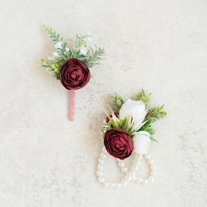 Könnte beinhalten: Zwei Ansteckblumen in Burgunderrot und Weiß mit Perlenakzenten. Die Ansteckblume links hat eine einzelne burgunderrote Blume und Grün. Die Ansteckblume rechts hat zwei weiße Blumen und eine burgunderrote Blume mit Grün und einer Perlenkette.