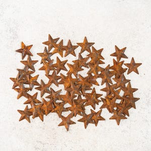 May include: A collection of small, rustic metal stars with a weathered, orange-brown finish. The stars are scattered on a light gray, textured surface, creating a decorative arrangement. Ideal for crafting or home decor.