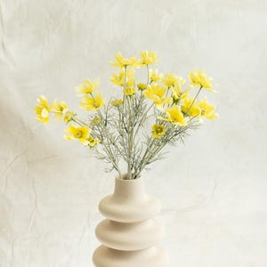 May include: A decorative vase in a cream colour, with a stacked, rounded design, holds a vibrant bouquet of yellow flowers. The flowers have delicate green stems. The background is a soft, off-white fabric, creating a simple, elegant composition.