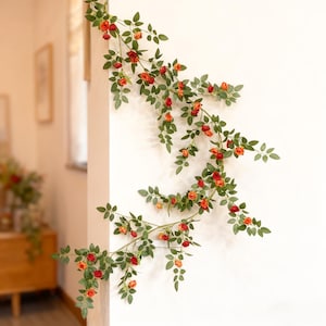 May include: Artificial rose garland featuring red and orange flowers and green foliage. The garland is arranged on a white wall, forming a decorative floral arrangement. The roses are in different stages of bloom.