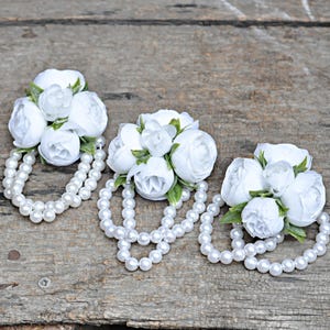 Puede incluir: Tres pulseras de corsage floral blanco con pulseras de perlas. Cada corsage presenta un grupo de flores de tela blanca con hojas verdes. Las pulseras de perlas están hechas de pequeñas perlas blancas redondas. El fondo es una superficie de madera envejecida.