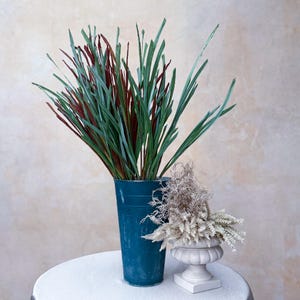 Puede incluir: Un jarrón de metal color turquesa lleno de tallos verdes y burdeos parecidos a hierba. A la derecha, una pequeña urna blanca contiene plantas secas de color beige. El arreglo está sobre una superficie blanca sobre un fondo neutro.