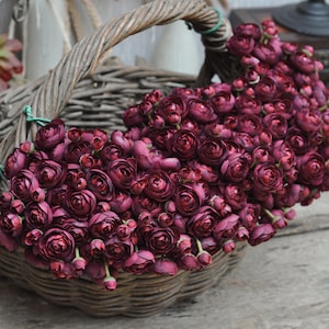 May include: A wicker basket filled with deep burgundy artificial flowers. The flowers are densely packed, with numerous buds and open blossoms. The basket features a curved handle and is set against a neutral backdrop, creating a rustic, natural aesthetic.