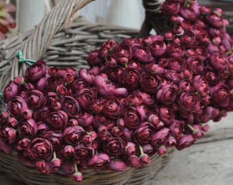 Pode incluir: Uma cesta de vime tecida transbordando de flores artificiais de cor bordô. As flores estão densamente agrupadas, com muitos botões e flores abertas. A cesta tem uma alça curva e está colocada sobre um fundo neutro, criando uma estética rústica e natural.