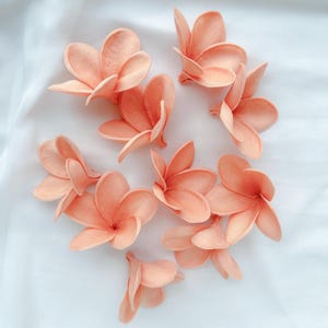 May include: A collection of peach-colored artificial plumeria flowers, arranged on a white surface. The flowers have multiple petals and a realistic appearance, suitable for crafting or decorative purposes.