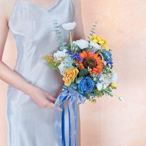 May include: A vibrant bouquet with an orange sunflower, blue roses, and yellow and white blooms. The bouquet is tied with a blue ribbon. The flowers are held by a person wearing a silver dress.