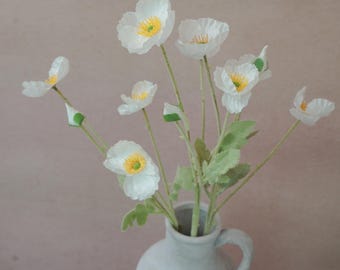 May include: A bouquet of white poppy flowers with yellow centers and green stems in a light gray ceramic vase. The flowers are in full bloom, with delicate petals and a natural appearance. The vase has a handle and a wide opening.