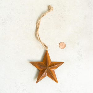 May include: A five-pointed star ornament made of rusted metal with a textured brown finish. The star hangs from a thin, light brown jute twine loop. A copper-colored penny is placed next to the star to provide a sense of scale for the object.