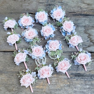 May include: A collection of floral boutonnieres featuring light pink roses, surrounded by blue and green accents. Each boutonniere has a pink stem. The flowers are arranged on a rustic wooden surface, creating a delicate and elegant display.