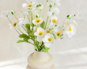 May include: A bouquet of white flowers with yellow centers and green leaves in a beige, stacked-ring vase. The flowers have delicate petals and are arranged to create a full, lush display. The vase sits on a white surface.