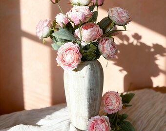 Pode incluir: Um buquê de peônias rosa em um vaso branco. As flores têm múltiplas camadas de pétalas e estão em vários estágios de floração, de botões a flores totalmente abertas. O vaso é de cor bege claro com uma superfície texturizada. O arranjo está sobre um tecido branco.