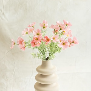 May include: A bouquet of pink cosmos flowers in a beige, stacked-ring vase. The flowers have delicate petals and yellow centers, with green stems and foliage. The vase is on a white surface against a white backdrop.