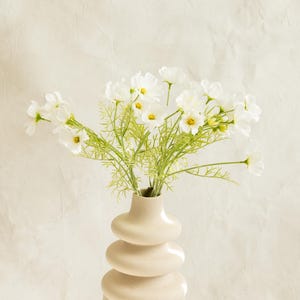 Könnte beinhalten: Eine beige Vase mit gestapelten Kugeln hält einen Strauß weißer Cosmosblüten mit gelben Zentren und grünem Laub. Die Vase steht auf einer weißen Oberfläche vor einem neutralen Hintergrund, ein minimalistisches Blumenarrangement.