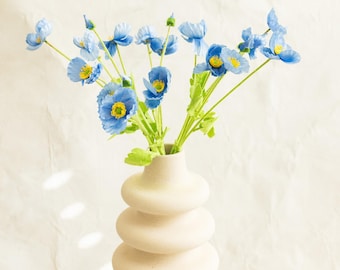 May include: A bouquet of blue poppy flowers with yellow centers and green stems in a cream-colored, stacked-disc vase. The flowers are in full bloom, creating a vibrant contrast against the neutral background.