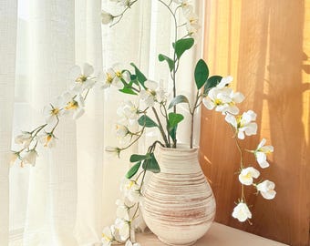 Puede incluir: Un jarrón de cerámica blanca con un diseño a rayas texturizado contiene flores blancas con hojas verdes. Las flores caen en cascada, creando una presentación natural y elegante. El jarrón está sobre una superficie blanca.