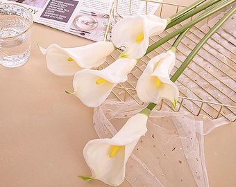 May include: Five artificial calla lilies with white petals and yellow centers are arranged on a gold wire basket. The stems are green. A sheer, star-patterned fabric and a glass of water are also visible.