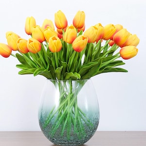 May include: A bouquet of yellow and orange artificial tulips in a clear glass vase with a textured surface.