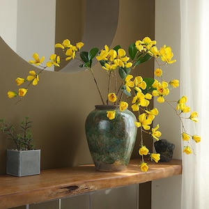 May include: A floral display with vibrant yellow flowers in a green and brown ceramic vase. The flowers are arranged with green leaves. A small potted plant and a dark stone are on a wooden shelf.